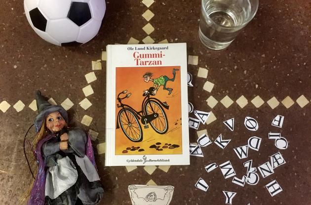 Bogen Gummi-Tarzan på et gulv omringet af bl.a. et glas vand, en fodbold og en heks