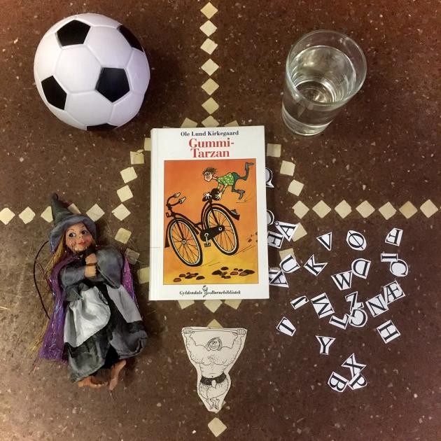 Bogen Gummi-Tarzan på et gulv omringet af bl.a. et glas vand, en fodbold og en heks