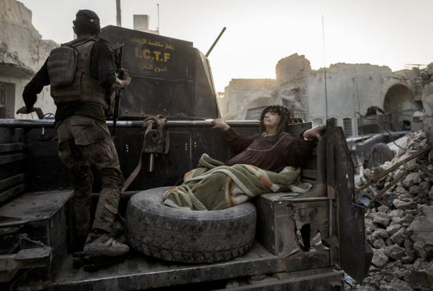 En soldat og en ældre mand er på laget af en vogn. Den ældre mand ligger ned med benene i et dæk. Området omkring dem er bombet.