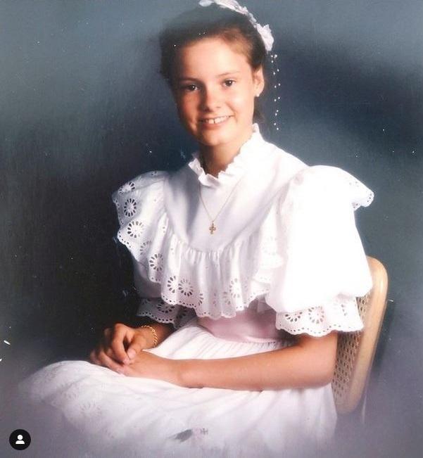 En ung Leonora Christina Skov sidder i en hvid kjole med opsat hår. Hun smiler. Baggrunden er mørk.